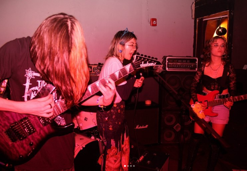ID: A pink and red tinted image of Cat Crash performing. Cecil Yang, an Asian-American person wearing a cat ear headband, stands in the center, singing into a microphone. To the left of them, Brett Cornball plays guitar, with their hair completely covering their face. To the right of Cecil, Gabrielle Blevins, a feminine person with curly brown hair, plays guitar and stares stoicly into the distance. END ID.