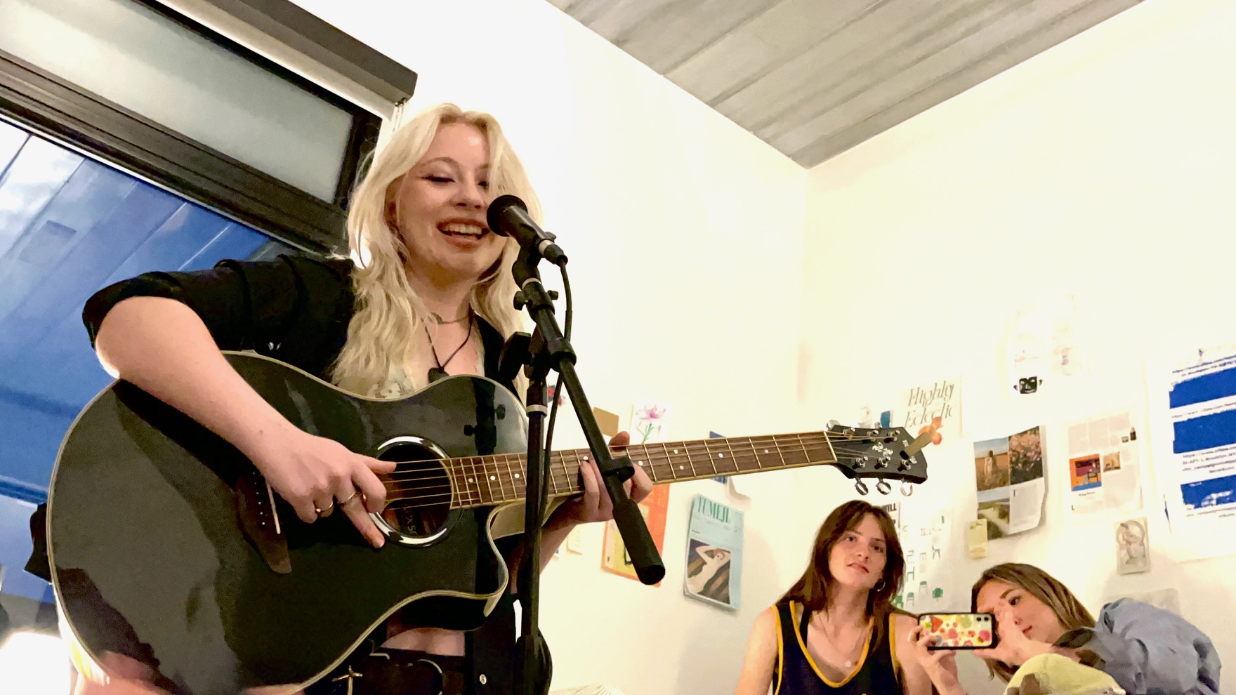 ID: Katie Overbey, a tall, white, feminine person with long blonde hair, sings into the microphone. She holds a black acoustic guitar. She is smiling. In the background, Zoe Stroupe sits next to Maya Ogea while she takes a picture on her phone. END ID.